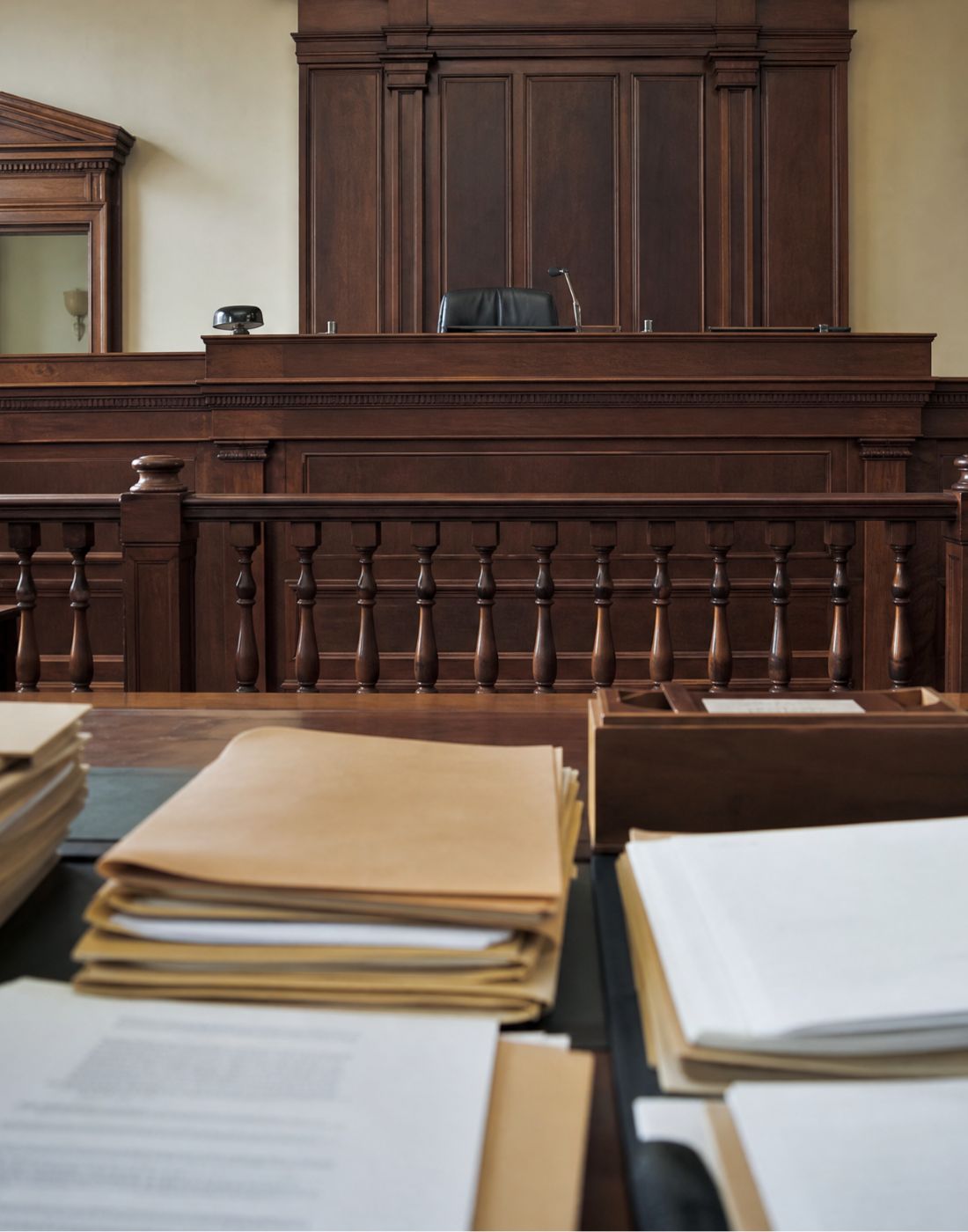 A court courtroom with a dark wood judges bench and paneled backdrop foregrounded by stacked legal folders and papers