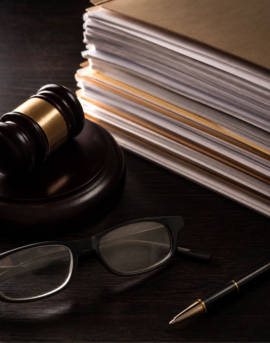 Judges gavel beside a thick stack of documents on a dark desk with glasses and a gold trimmed pen nearby